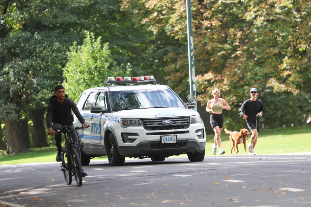 An NYPD police vehicle parked on a paved road in Central Park, with a cyclist passing in the foreground and a couple walking their dog nearby.