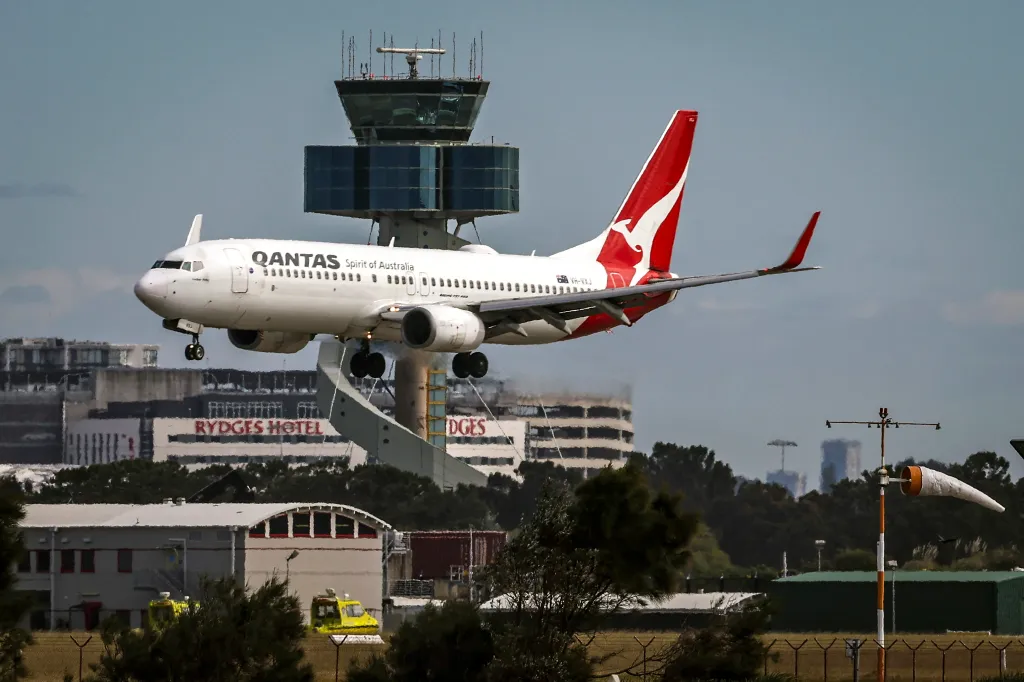 A Qantas Airways Boeing 737 aircraft approaches for landing in Sydney, with the airport's air traffic control tower in the background.
