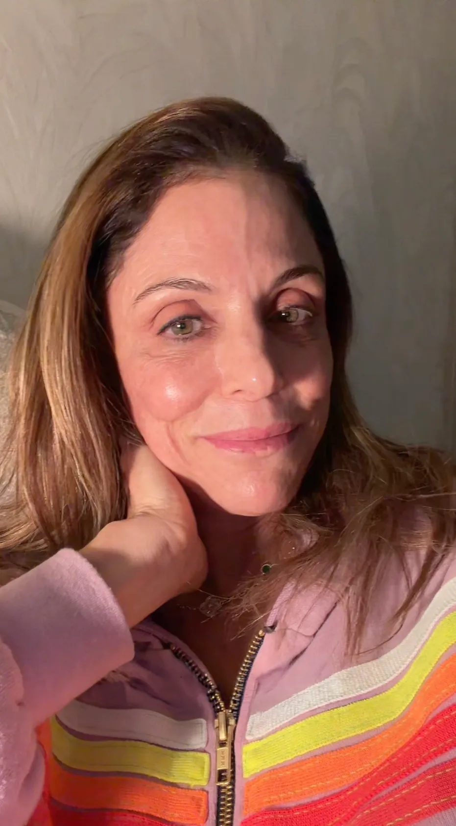 Bethenny Frankel wearing a pink zip-up hoodie with rainbow stripes, resting her hand on her neck.