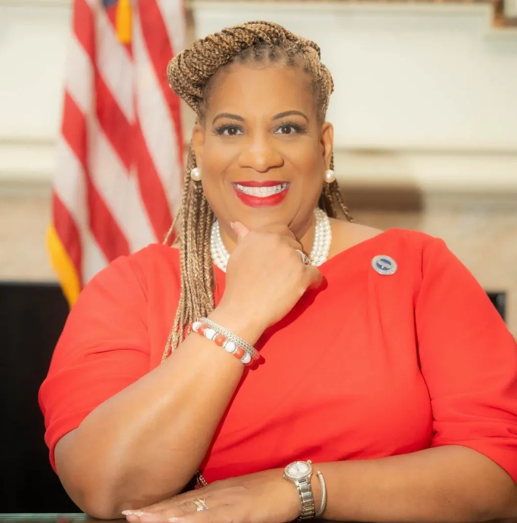 Shawyn Patterson-Howard smiling in a red dress with an American flag in the background.