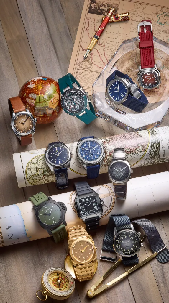 An arrangement of luxury watches laid out on a wooden surface alongside travel-themed items like a globe, maps, a compass, and a pen.