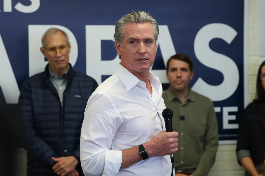 California Governor Gavin Newsom speaking into a microphone with two men standing behind him.