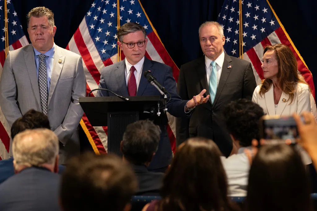 Mike Johnson speaking at a podium with Nick LaLota, Steve Scalise, and Lisa McClain standing behind him in front of American flags.