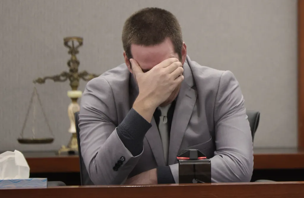 A man in a gray suit sits with his head in his hand, looking distressed in a courtroom setting.