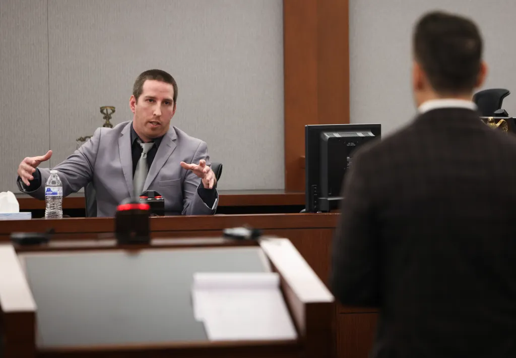 A man in a gray suit speaks in a courtroom, gesturing with his hands while looking toward someone standing in the foreground.