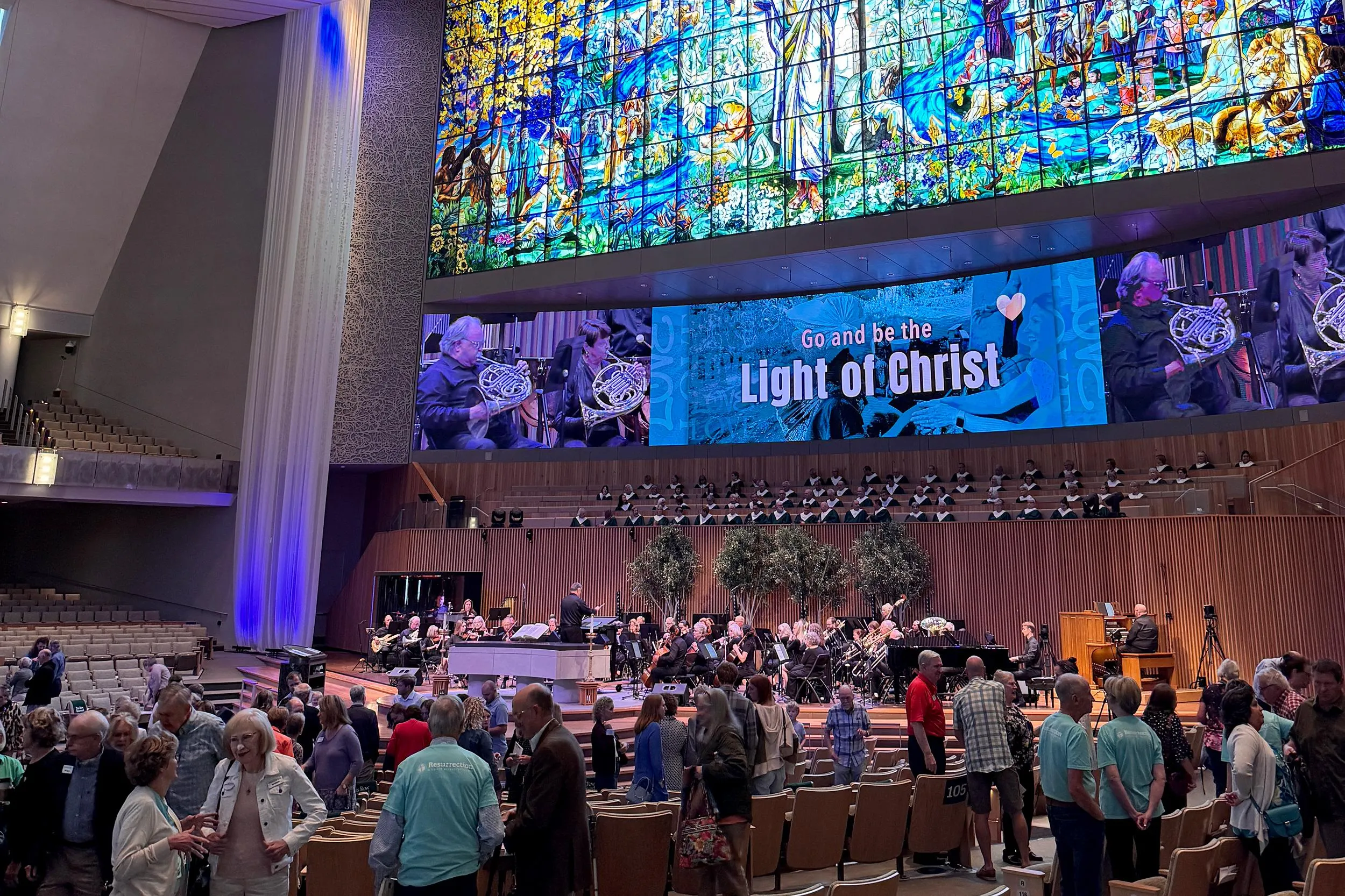 a wide view of the interior of a large church filled with people greeting one another. Light is streaming through ornate stained glass. A screen above a live orchestra reads “Go and be the Light of Christ” a wide view of the interior of a large church filled with people greeting one another. Light is streaming through ornate stained glass. A screen above a live orchestra reads “Go and be the Light of Christ”