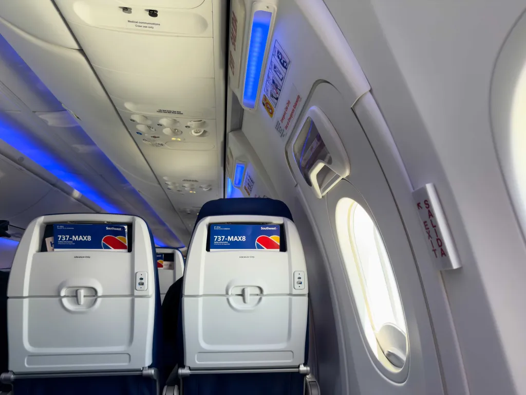 Interior view of the exit row of a Southwest Airlines Boeing 737 MAX 8 cabin, showing seats with 737-MAX8 headrests, overhead panels, and emergency instructions.