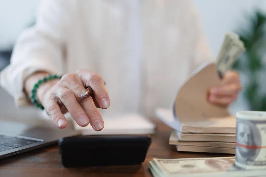 A senior woman calculates home finances, holding dollar banknotes and using a calculator, notebook, and laptop.