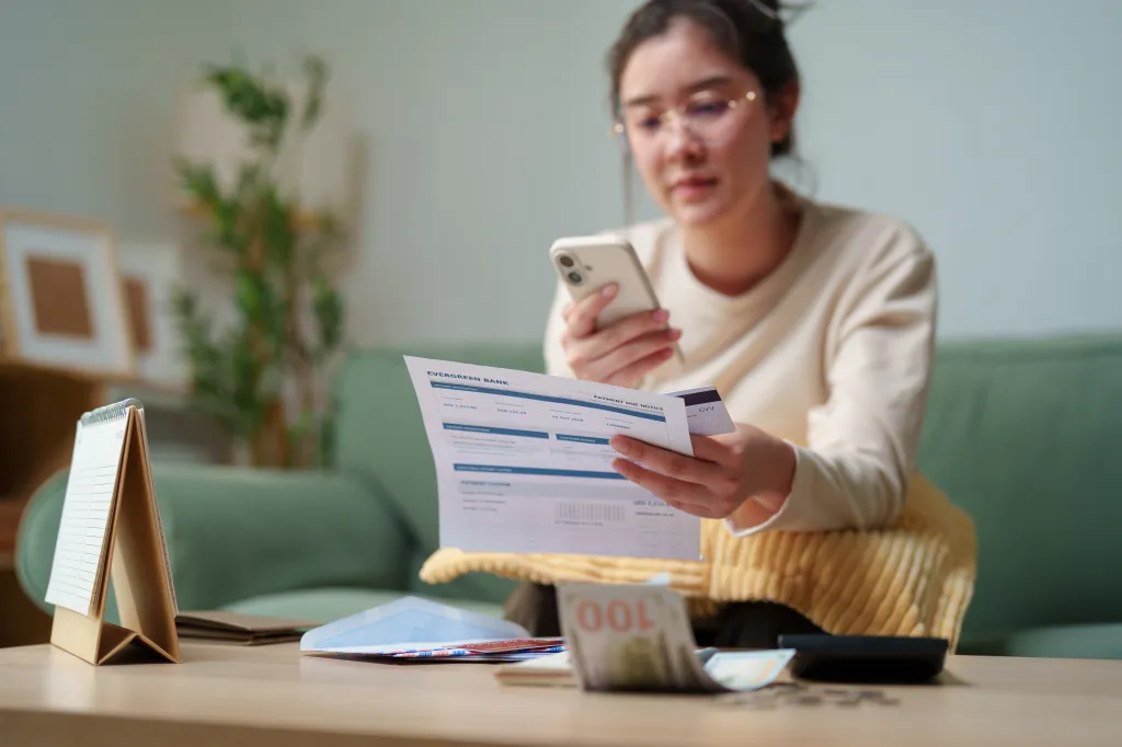 A woman checking her bank statement and holding a credit card and smartphone while managing her monthly payments at home.