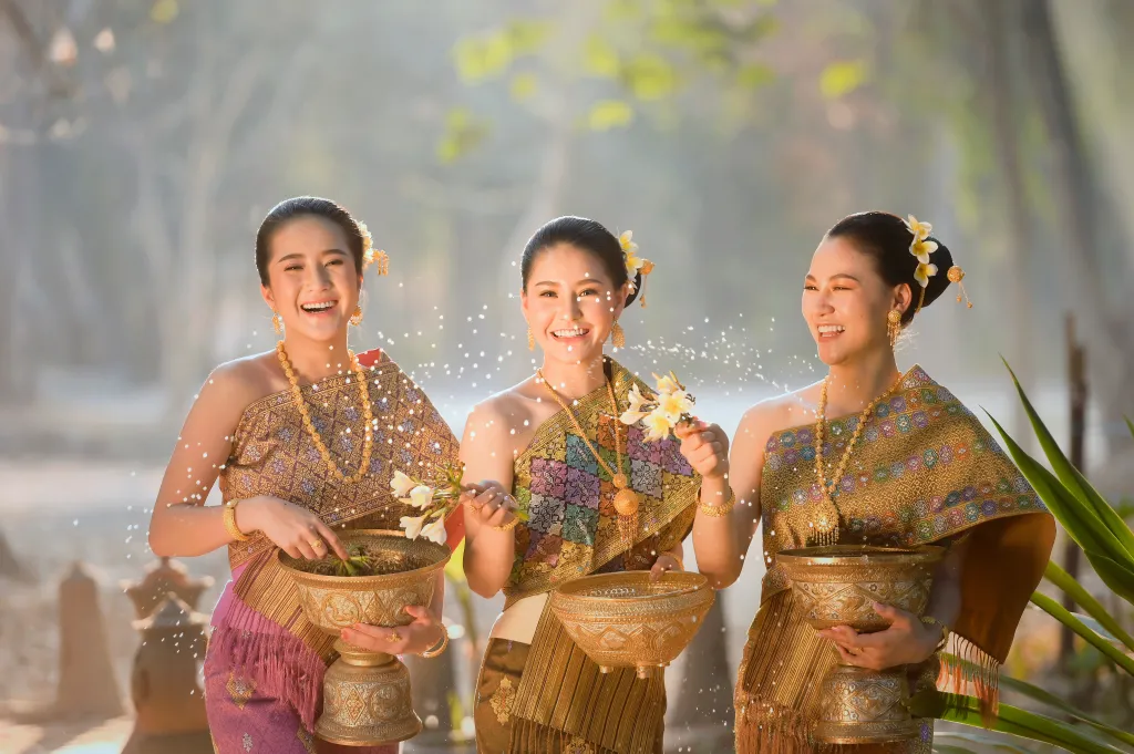 Three smiling women in traditional Thai attire celebrate the Songkran festival by splashing water and holding ornate bowls.