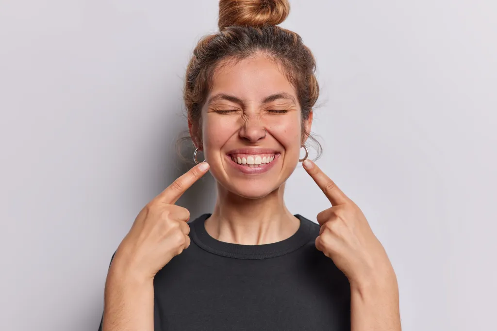 A cheerful woman wearing a black t-shirt points index fingers at her wide, toothy smile.