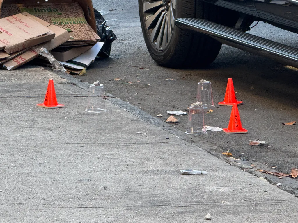 Crime scene markers, including orange cones and plastic cups weighted with stones, on a street near a vehicle and cardboard boxes.