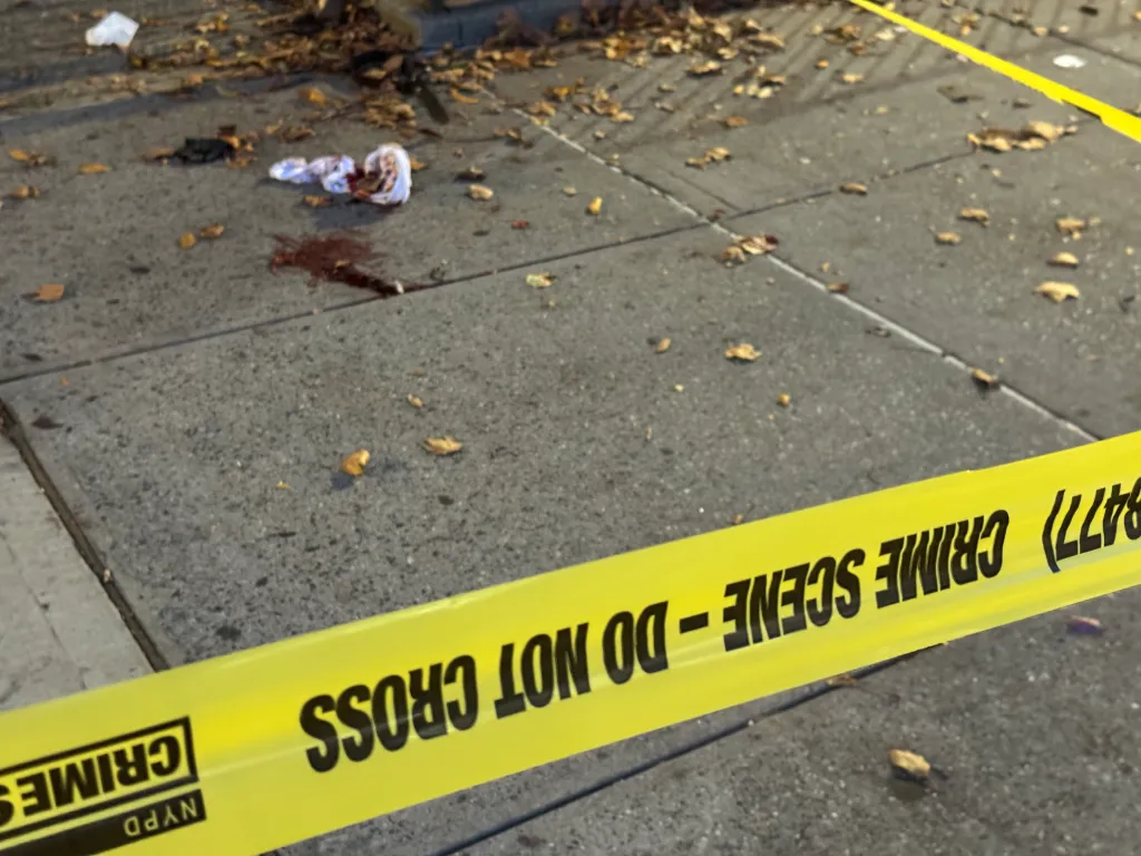 Yellow NYPD crime scene tape blocking off a sidewalk with blood and discarded material.