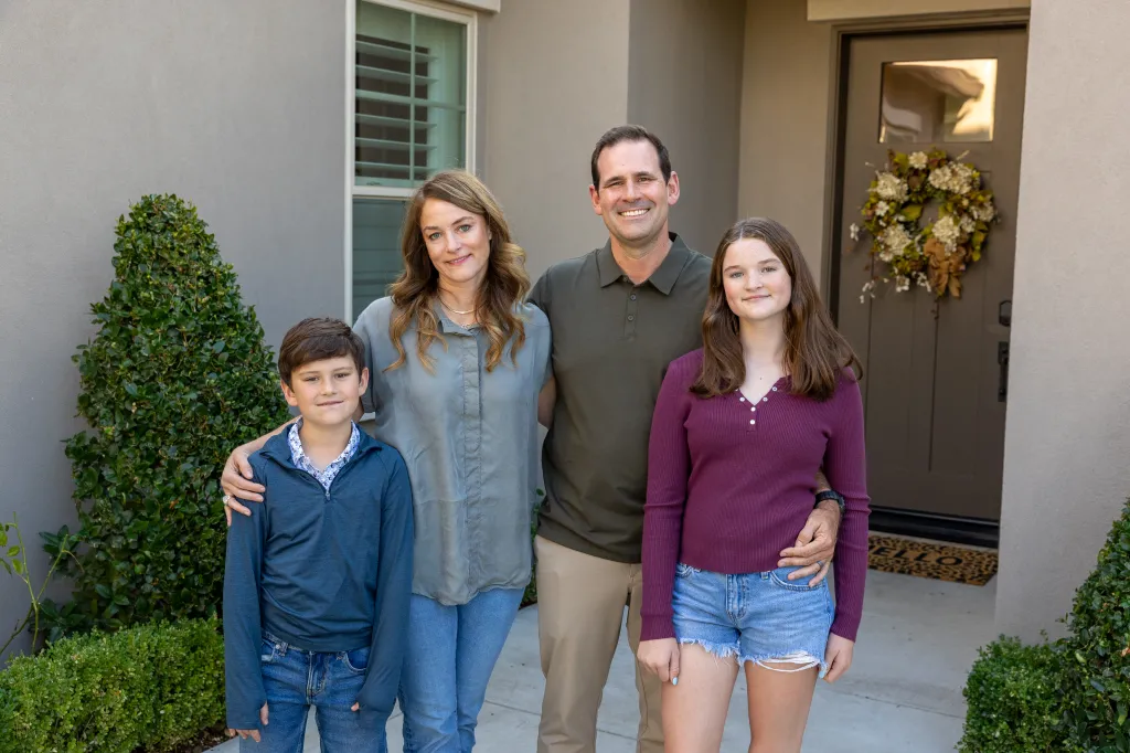 A family of four stands smiling in front of their home's entrance, with the parents in the middle and their son and daughter on either side.