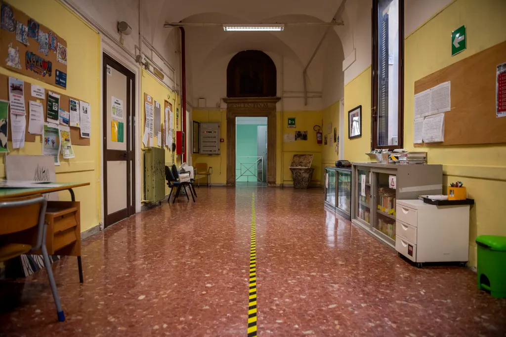 A school hallway with yellow walls, a tiled floor with a yellow and black safety line, and doors and bulletin boards along the sides.