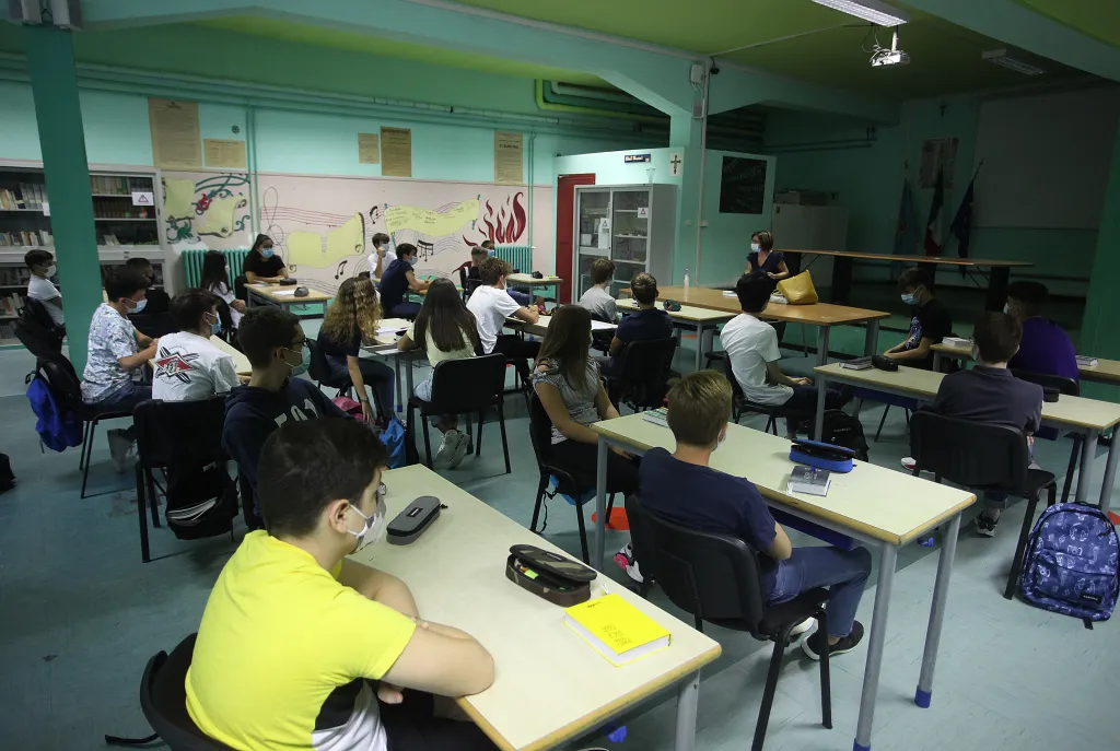 Students wearing face masks sit at desks in a socially distanced classroom while listening to a teacher.