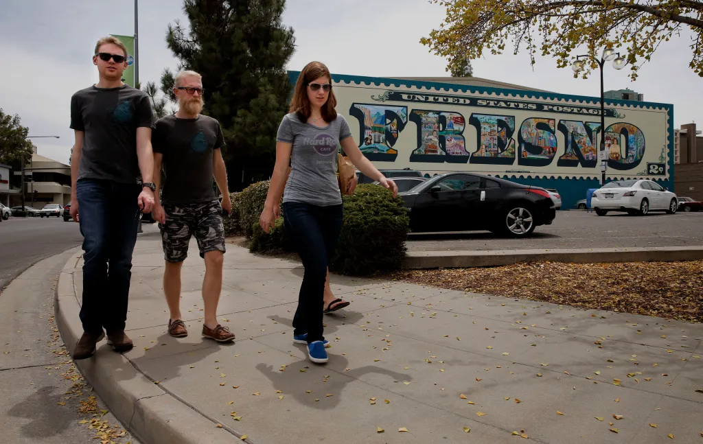 Three people walking on a sidewalk past a Fresno mural.