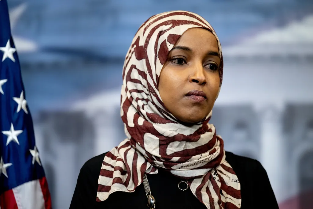 Representative Ilhan Omar wearing a patterned hijab, with an American flag visible in the background.