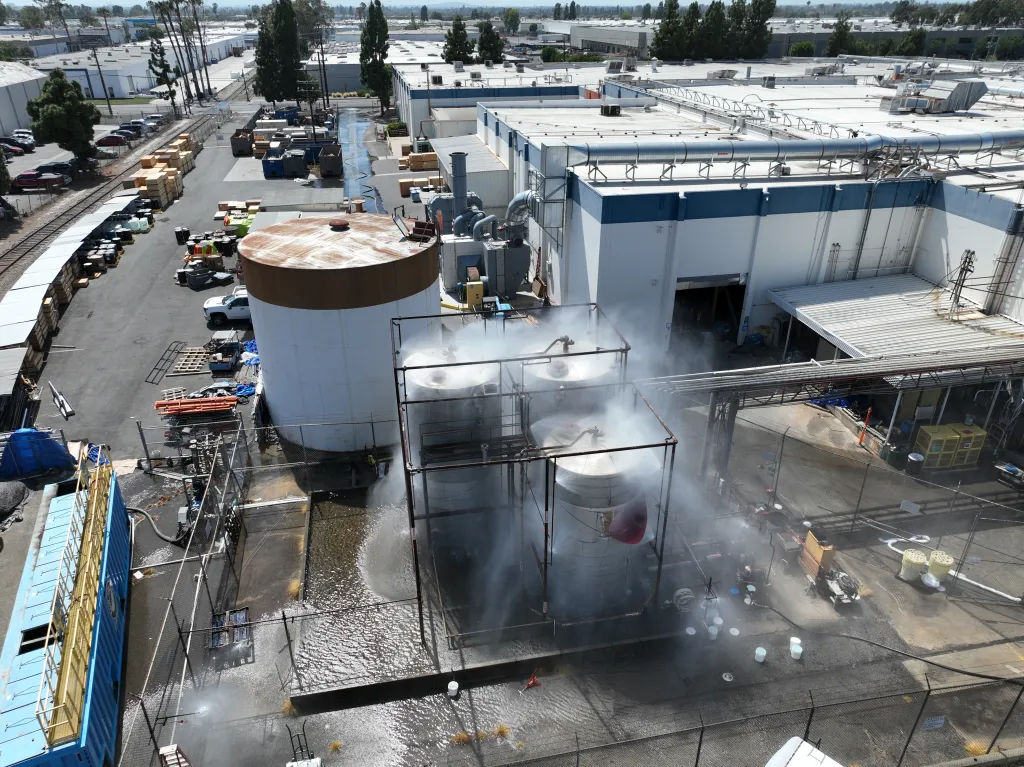 An aerial view of an industrial facility shows water spraying from a group of metal tanks surrounded by a metal cage, with a large storage tank nearby and factory buildings in the background.