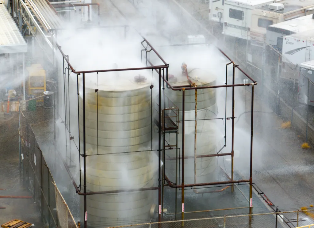 High-angle view of two large industrial tanks being sprayed with water from a surrounding pipe framework, with thick mist and steam filling the air.