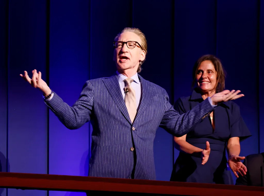 Bill Maher speaks at the 27th Mark Twain Prize For American Humor Gala, with a woman smiling behind him.