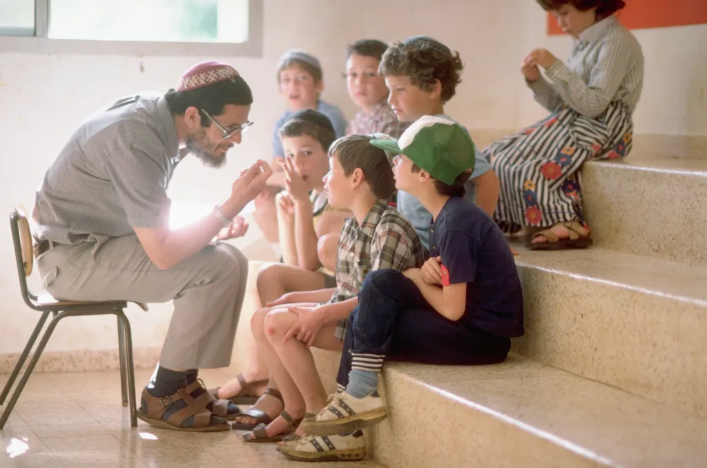 A teacher wearing a kippah sits on a chair, talking to a group of young boys sitting on a staircase.