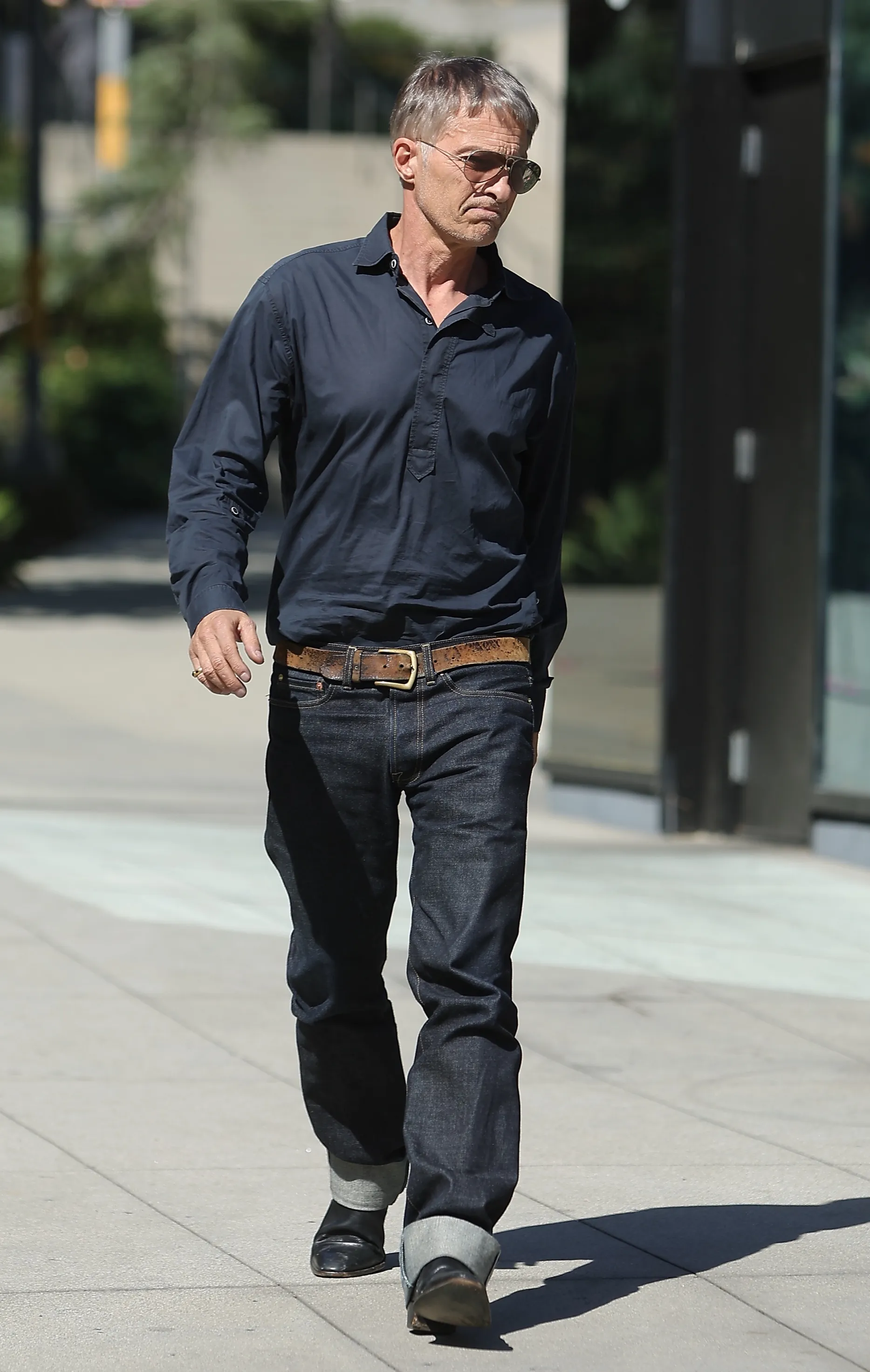 Billy Bob Thornton walking down a sidewalk, wearing sunglasses, a dark long-sleeved shirt, jeans, and a brown belt.