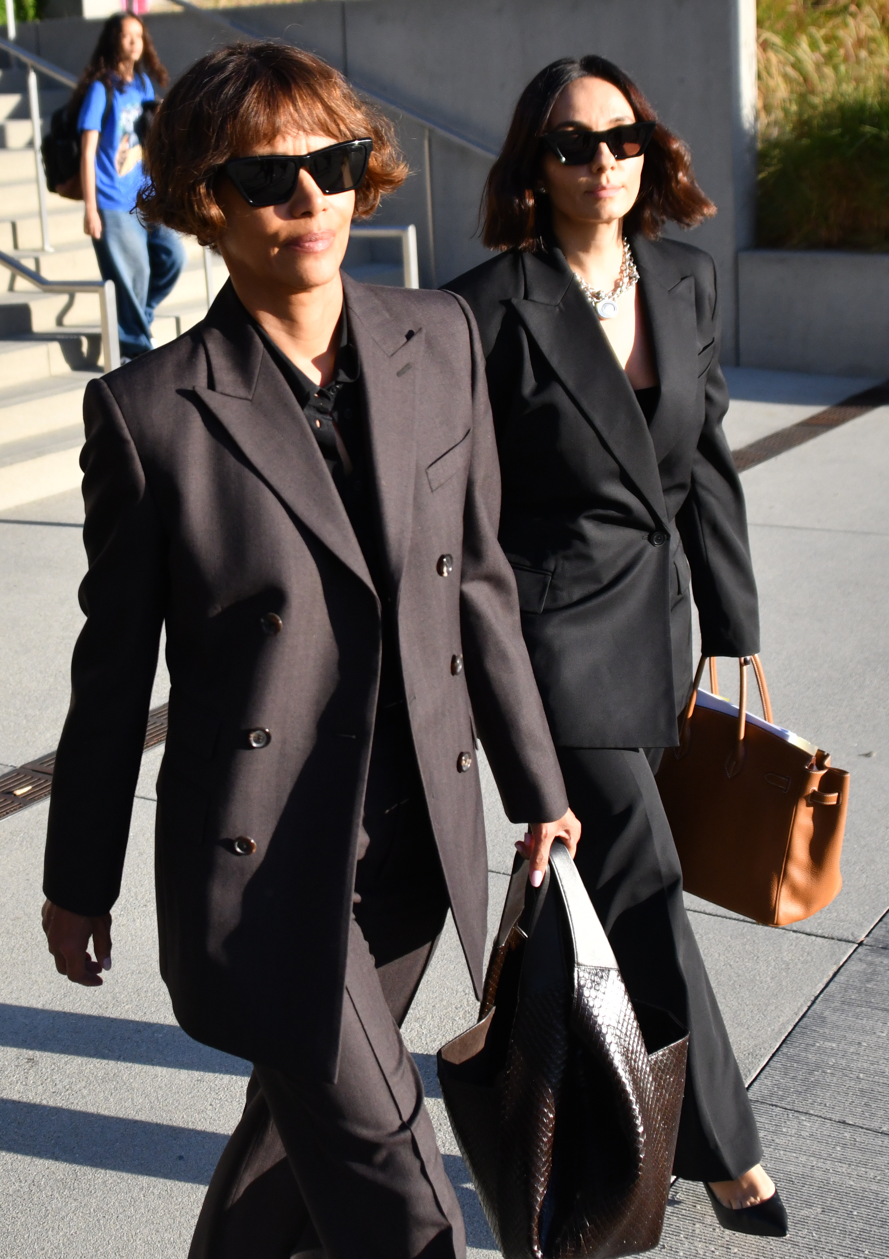 Halle Berry and a companion walking outside outdoors, both wearing black suits and sunglasses.