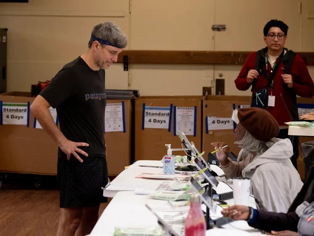 A voter in athletic wear stands at a polling table, talking to a poll worker in a brown beret inside a polling place.