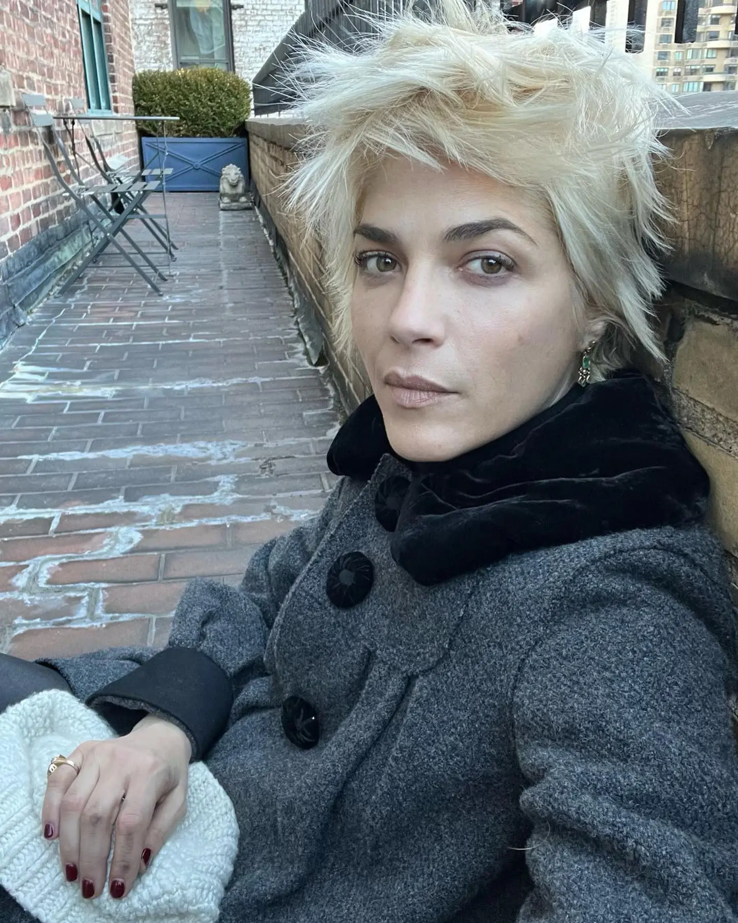 Selma Blair with short blonde hair on a brick balcony, wearing a grey winter coat.