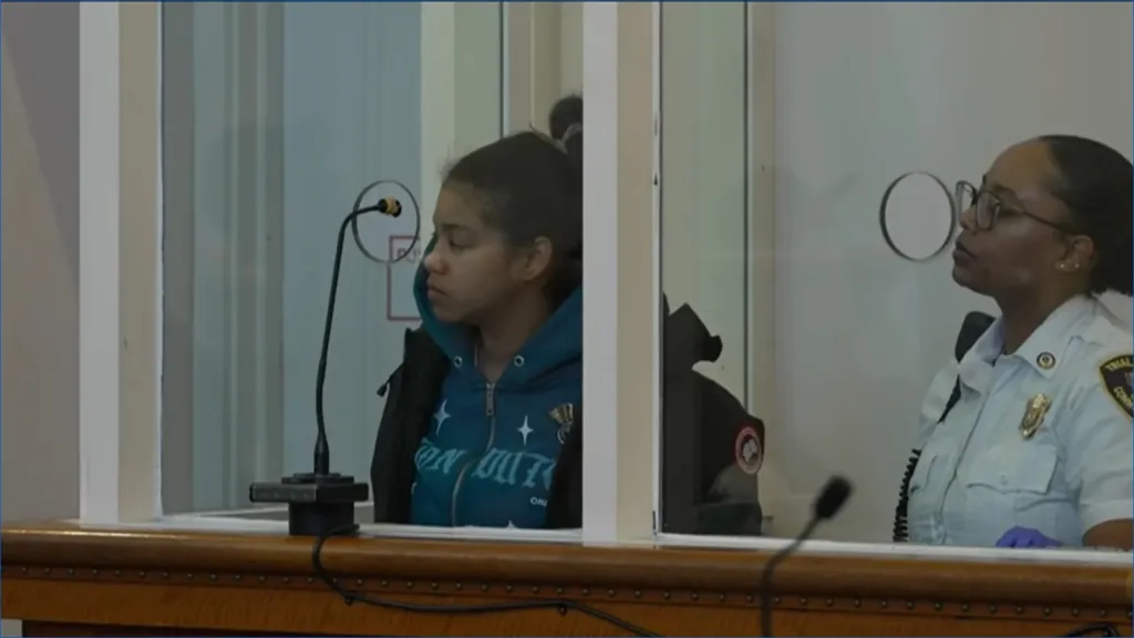 A woman sits in a courtroom behind a glass partition, with a female officer seated beside her.