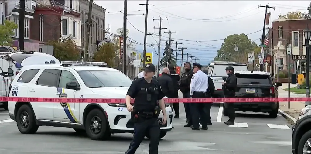Police officers standing on a city street behind red crime scene tape, near police vehicles and buildings.