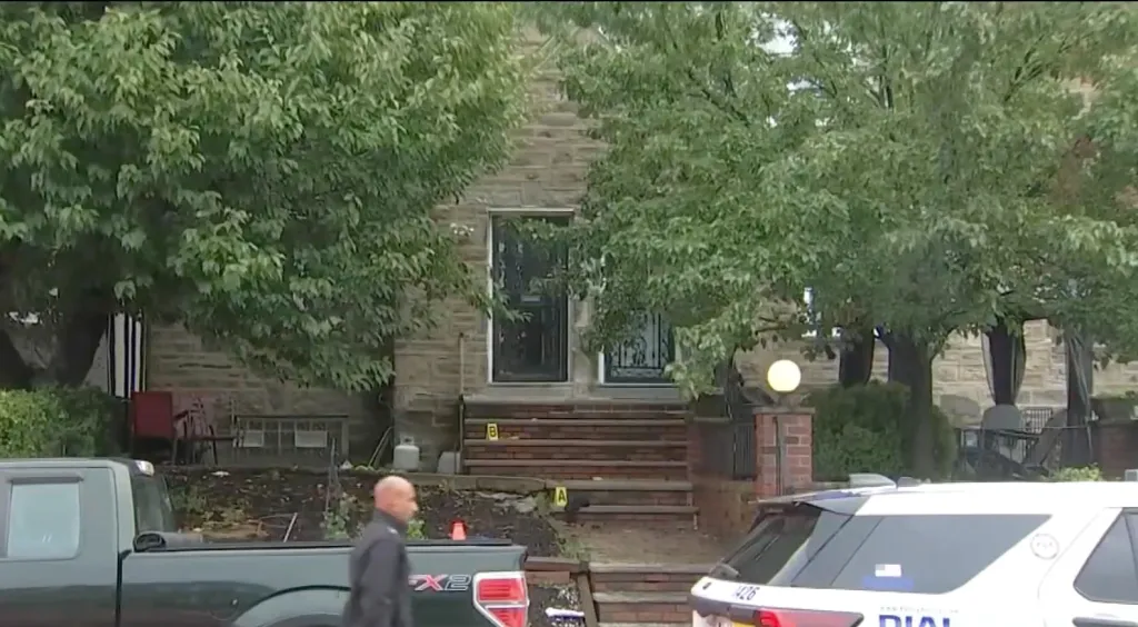 A person walks past police vehicles parked in front of a stone house with leafy trees.
