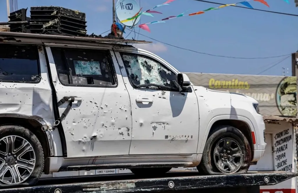 A white SUV riddled with bullet holes and with shattered windows, loaded onto a flatbed tow truck.