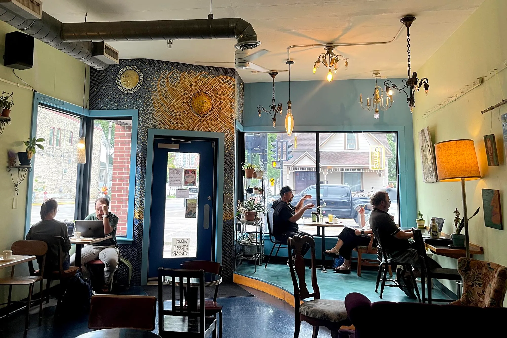 a blue, green, and yellow cafe interior with mosaic art and eclectic hanging lights. Several people are eating and working at small tables.