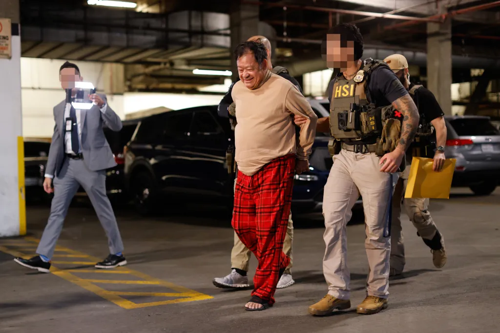 An older man in a beige sweater and red plaid pajama pants is escorted by law enforcement officers in a parking garage.
