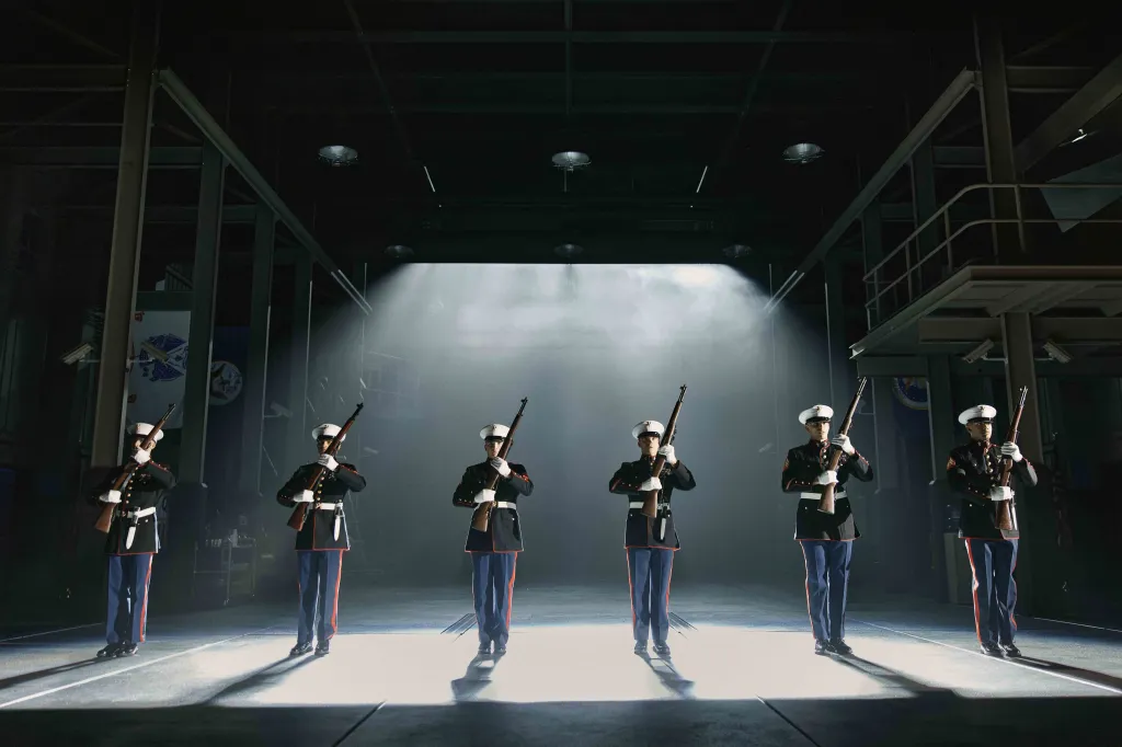 Six Marines in dress blues stand in a line on a stage, holding rifles and facing forward in a beam of spotlight.