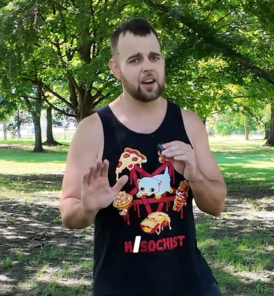 A man in a black tank top with a cartoon design stands in a park with trees.