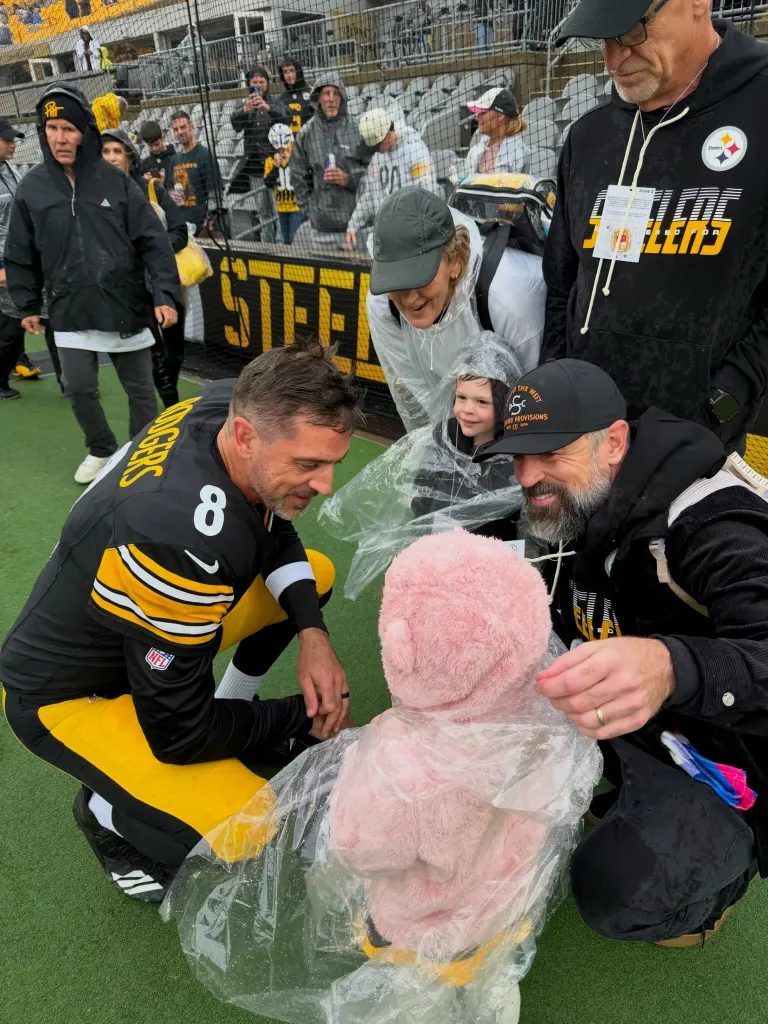 Aaron Rodgers and his family pose with a child in a pink bear suit covered in a plastic poncho on the sidelines of a Steelers game.