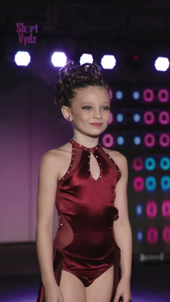 A young female dancer in a burgundy costume stands on stage under studio lights.