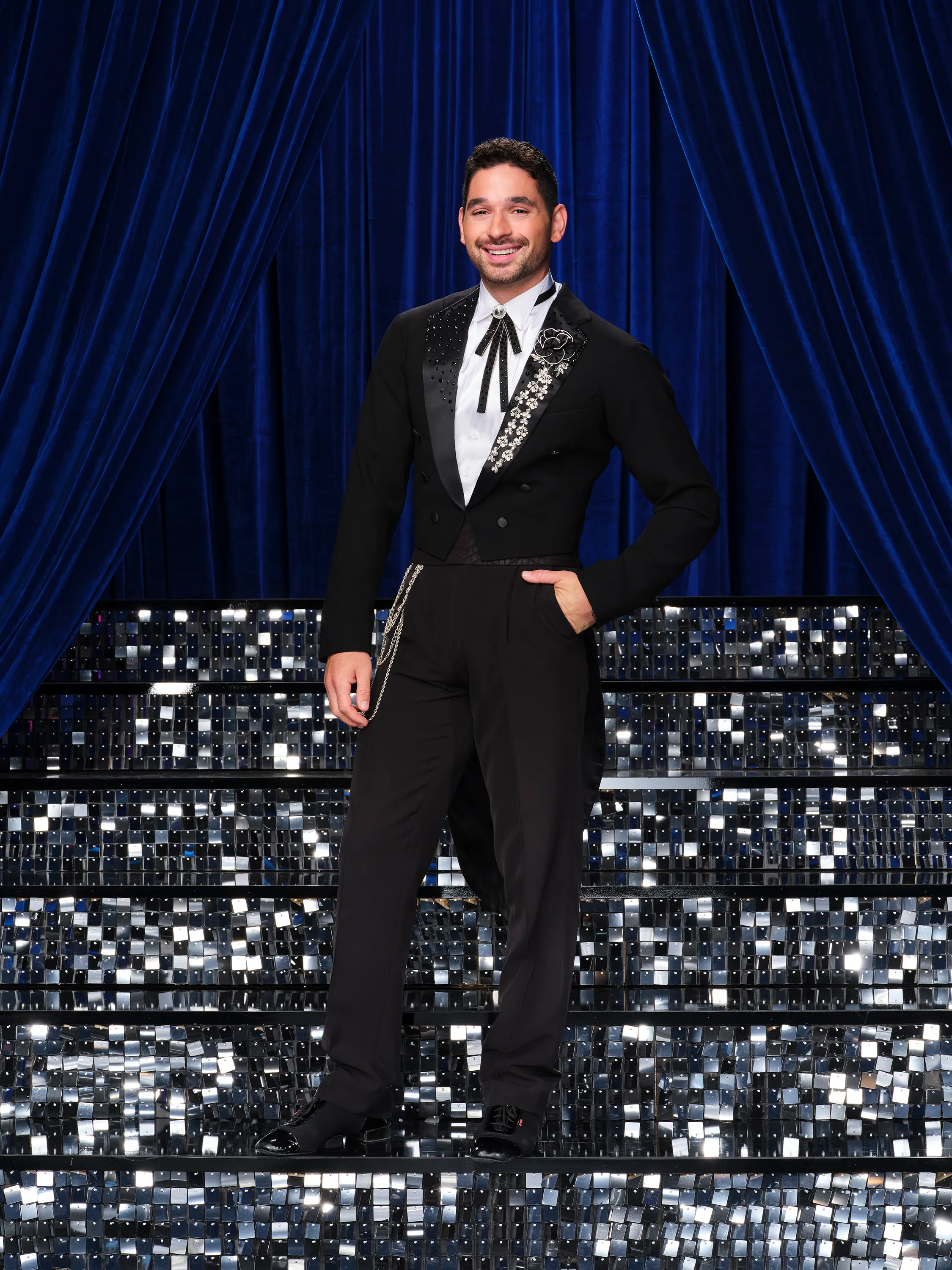 Alan Bersten poses in a black tuxedo on a glittering staircase in front of blue curtains.