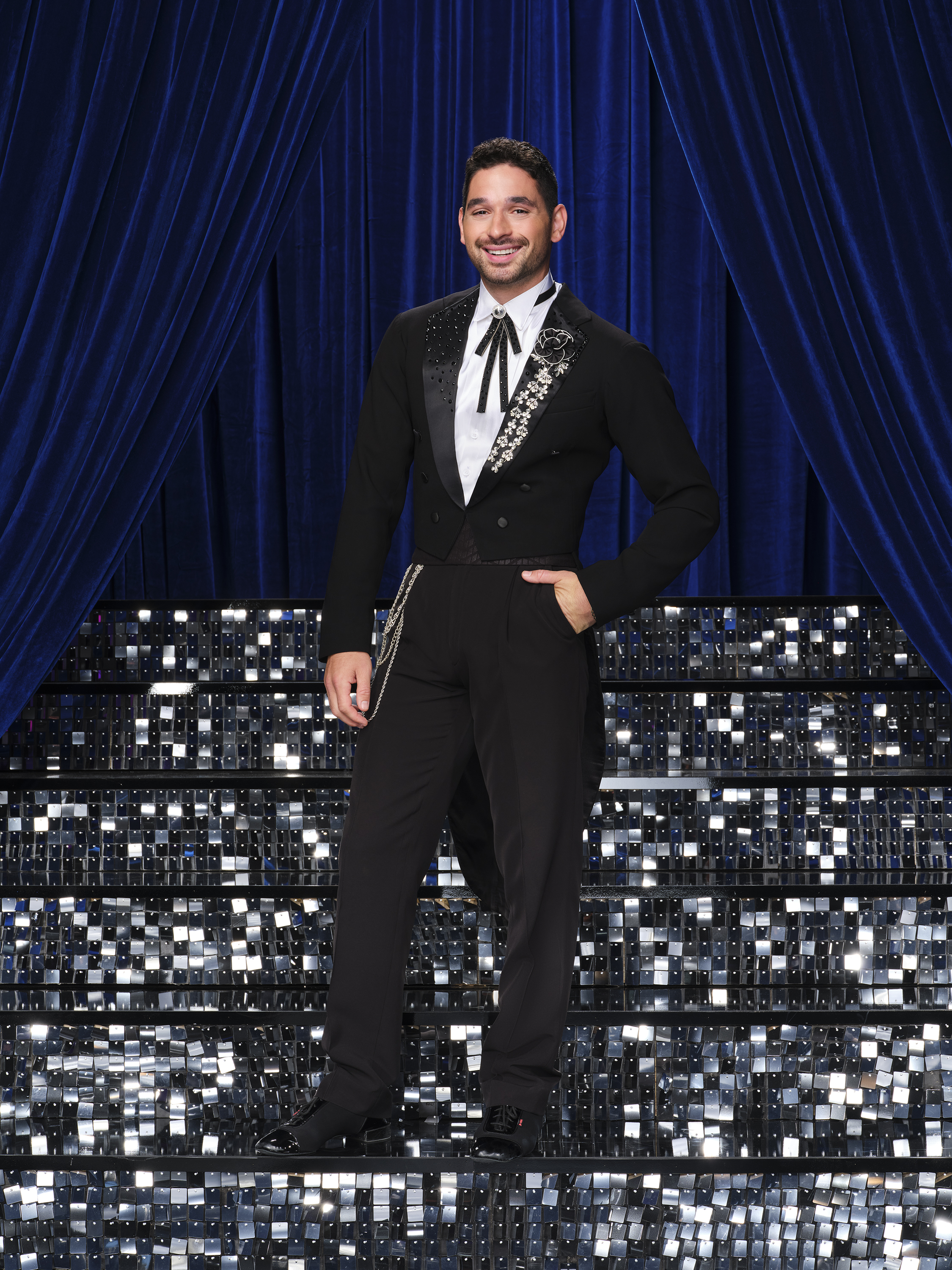 Alan Bersten poses in a black and white tuxedo in front of blue curtains and sparkly stairs.