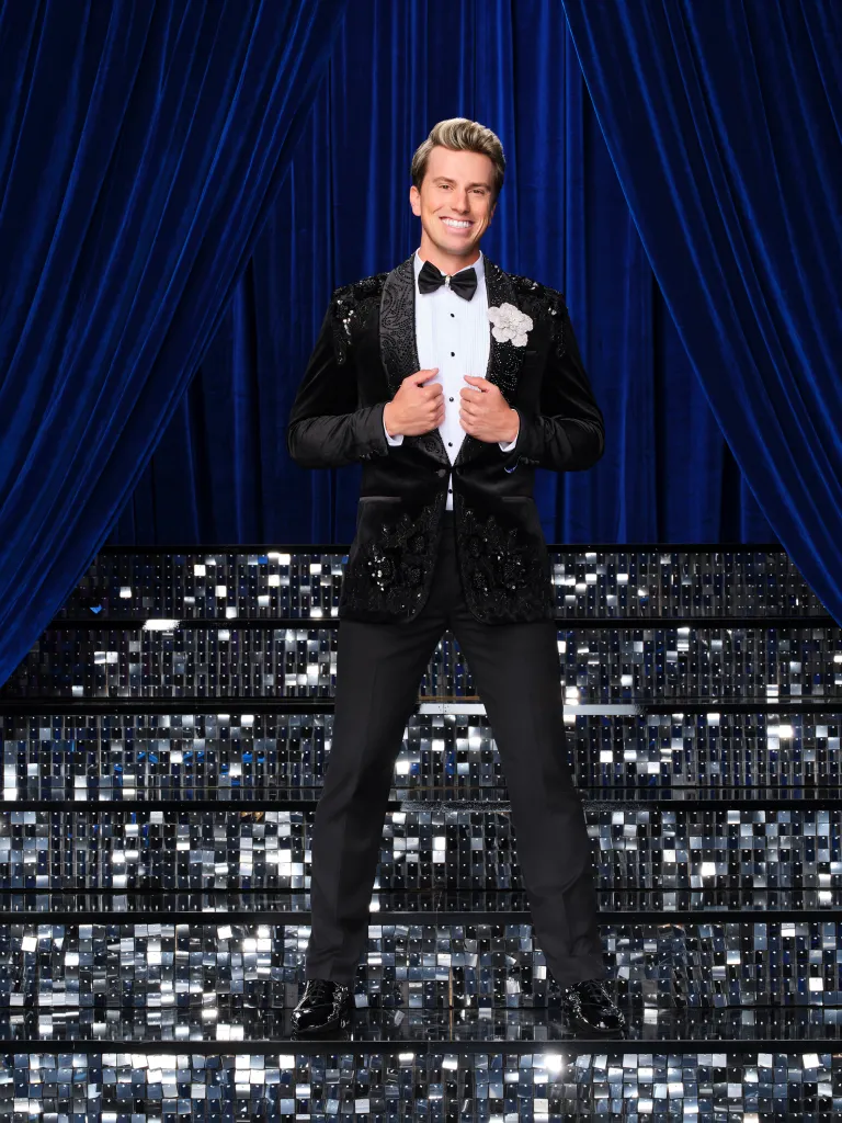 Jackson Olson stands on a sparkling stage in front of a blue curtain, wearing a black tuxedo with a white shirt and bowtie.