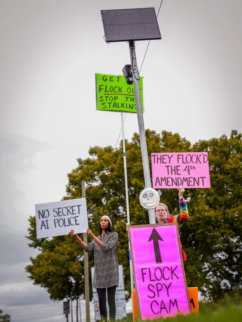 Activists with the Close the Camps Coalition stand beneath a Flock Safety camera holding protest signs that read 