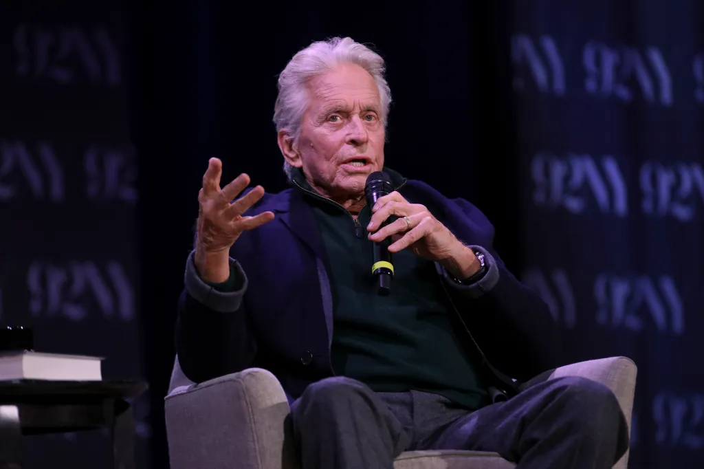 Actor Michael Douglas sitting in a chair, holding a microphone and gesturing while speaking on stage.