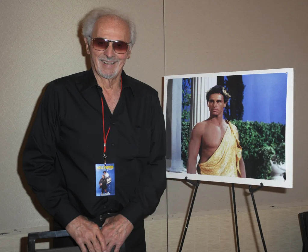 Actor Michael Forest posing next to a framed photo of himself in costume.
