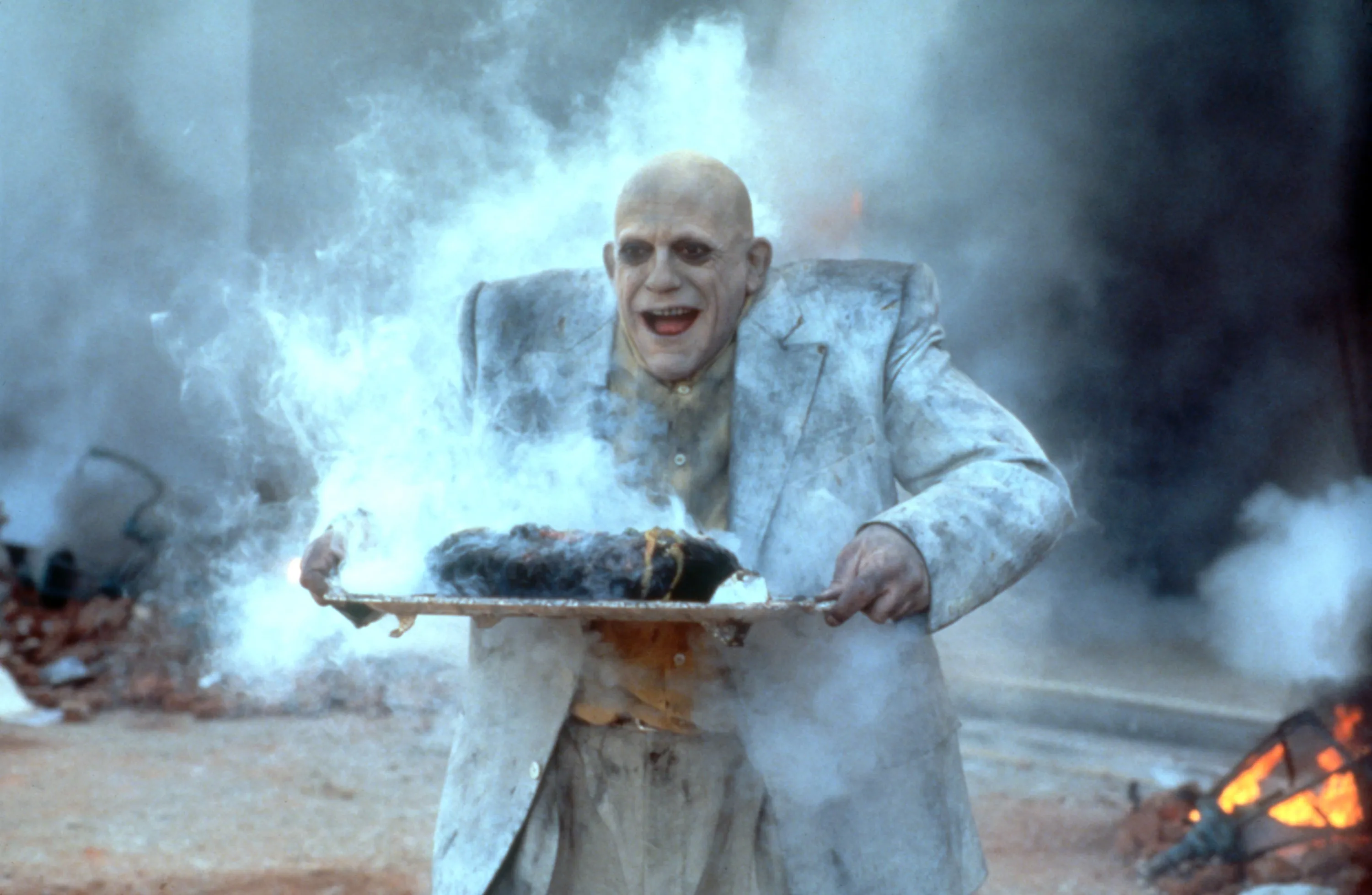Christopher Lloyd as Uncle Fester holding a smoking platter amidst smoke and debris.