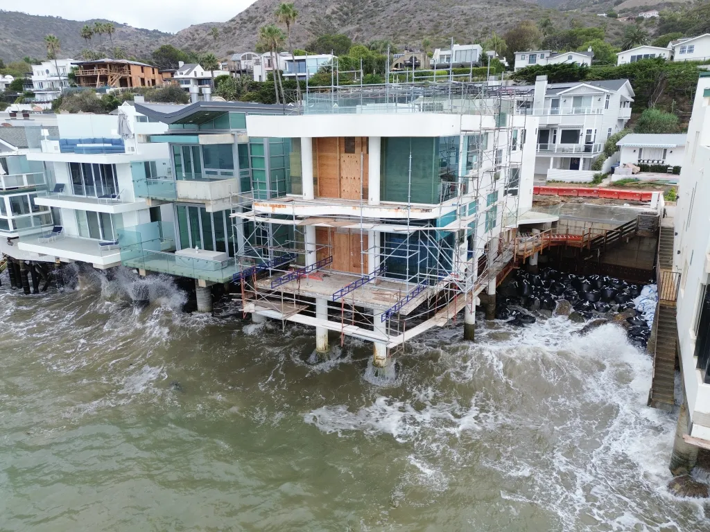 Aerial view of a beachfront house under construction with scaffolding, surrounded by rough, high-surf ocean water in Malibu, California.