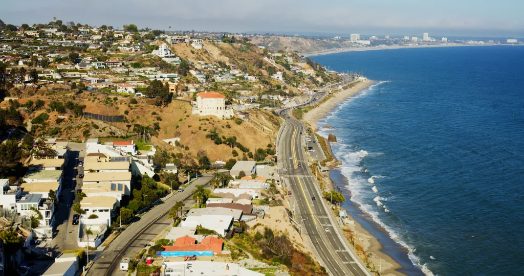 Aerial view of the Pacific Coast Highway running between the Pacific Palisades bluffs and the Santa Monica Bay shoreline.