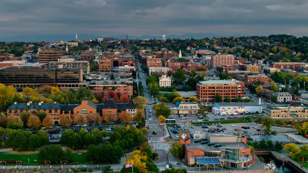 Aerial view of Burlington, Vermont, featuring historic buildings, trees with changing foliage under a hazy sky, and the Green Mountains in the distance.